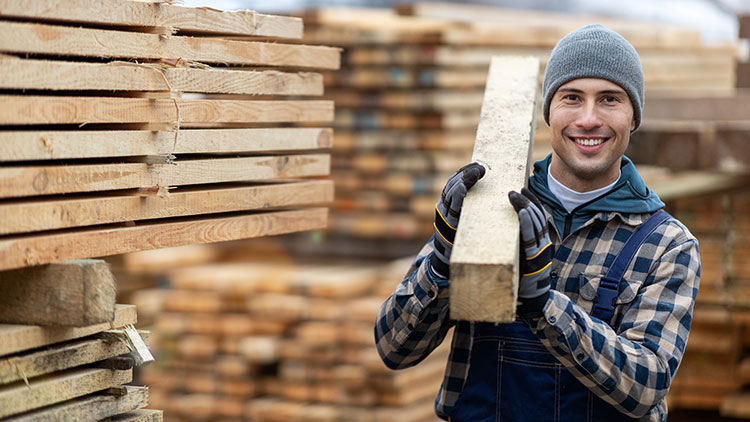 JUnger Mann trägt einen Holzbaklen in einem Holzlager