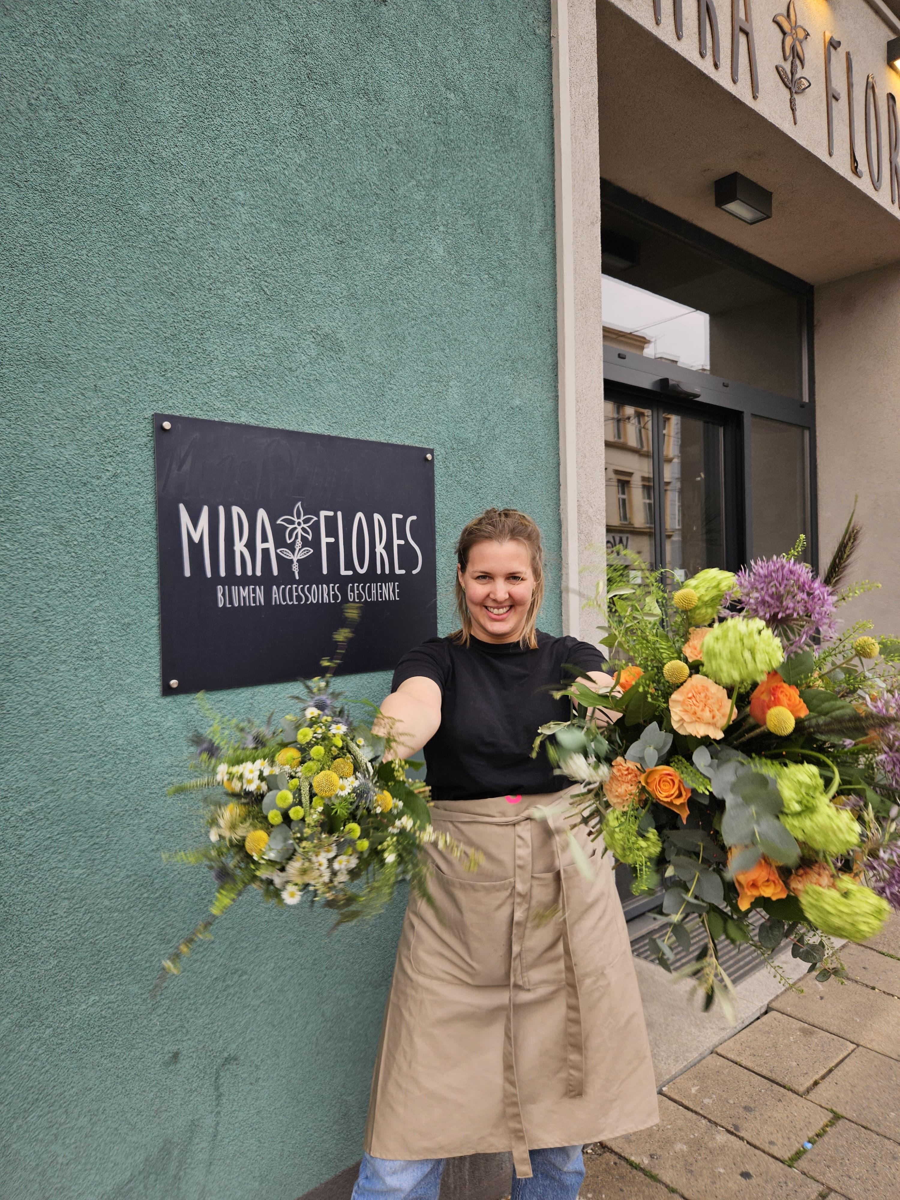 Floristin lächelnd mit Blumensträußen in beiden Händen vorm Geschäft