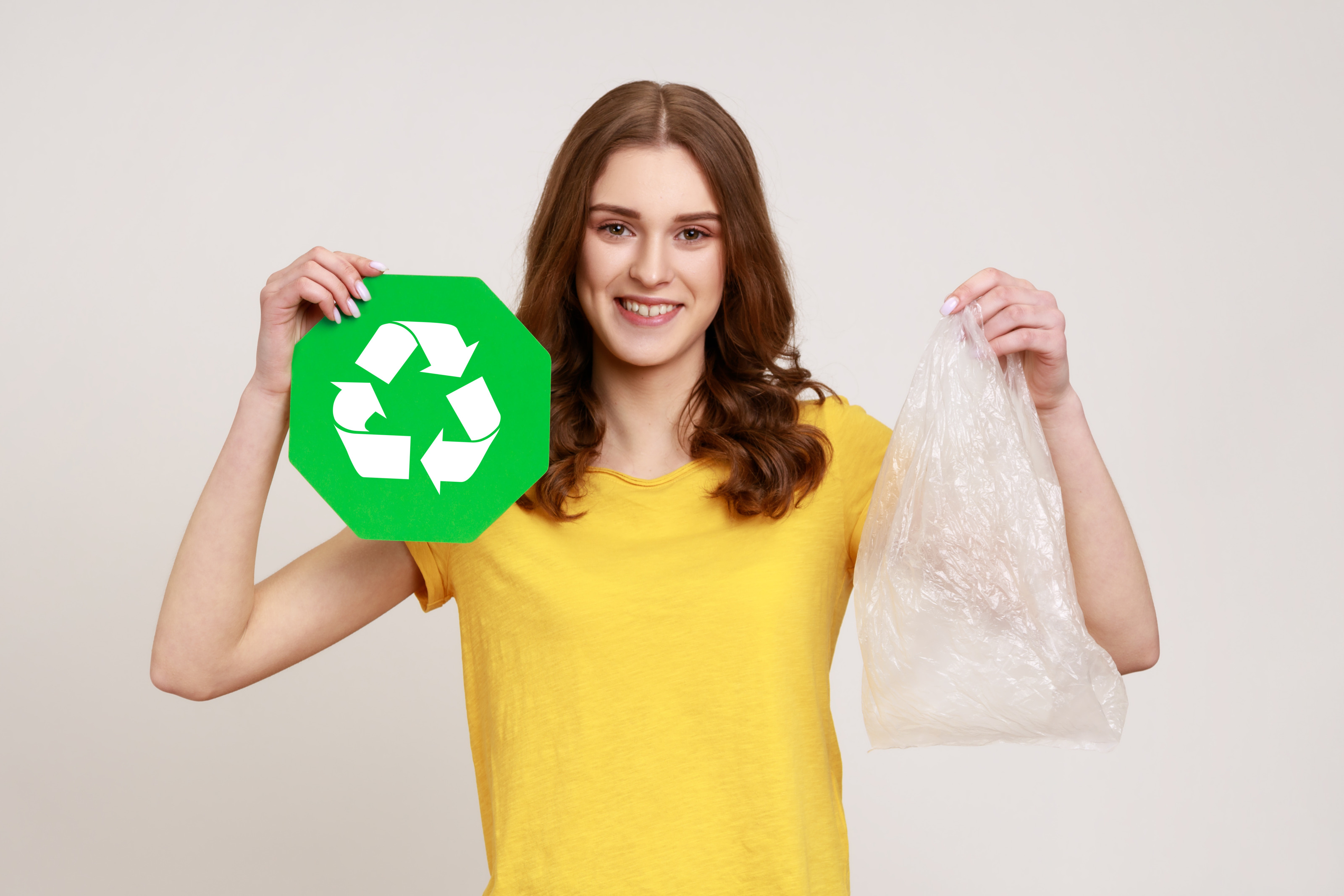 Portrait einer lächelnden Person in gelbem T-Shirt, die in einer Hand Recyclingzeichen - drei weiße im Kreis verlaufenden Pfeile auf weißem Hintergrund - und in anderer Hand Plastiksack hält